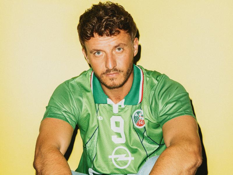Porträt von Tommi Schmitt mit lockigem Haar und Dreitagebart vor gelbem Hintergrund. Er trägt ein grünes Retro-Fußballtrikot mit der Nummer 9, dem Opel-Logo und dem Wappen des irischen Fußballverbands. Sein Blick ist ernst und direkt in die Kamera gerichtet.