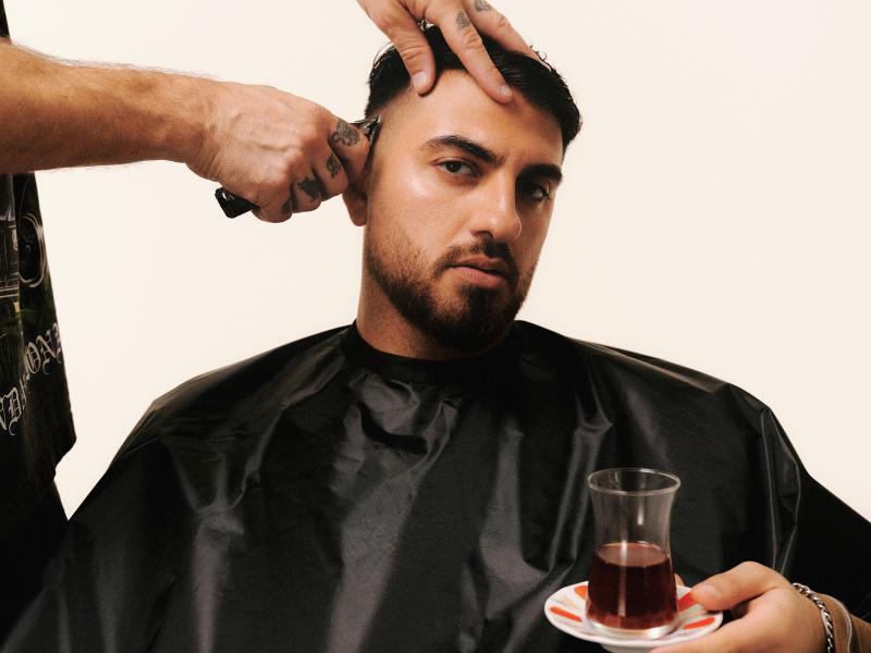Ein junger Mann mit Dreitagebart sitzt in einem schwarzen Friseurumhang und lässt sich die Haare schneiden. Eine tätowierte Hand führt die Haarschneidemaschine, eine andere reicht ihm ein Glas Tee. Sein Blick ist ruhig und leicht zur Seite gerichtet.