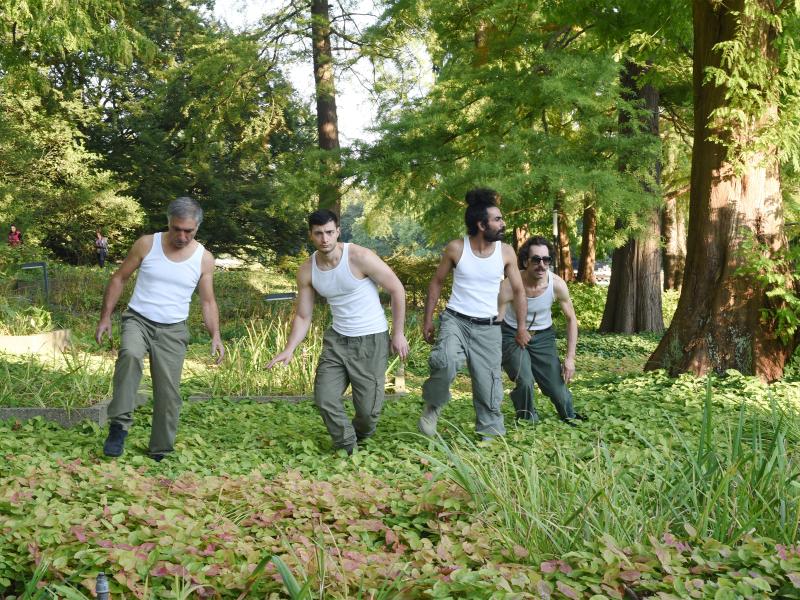 Vier Männer in weißen Tanktops und grauen Hosen bewegen sich durch einen dicht bewachsenen Park. Im Hintergrund stehen große Bäume und weitere Spaziergänger; die Männer sind von üppiger Vegetation und Rasenflächen umgeben.