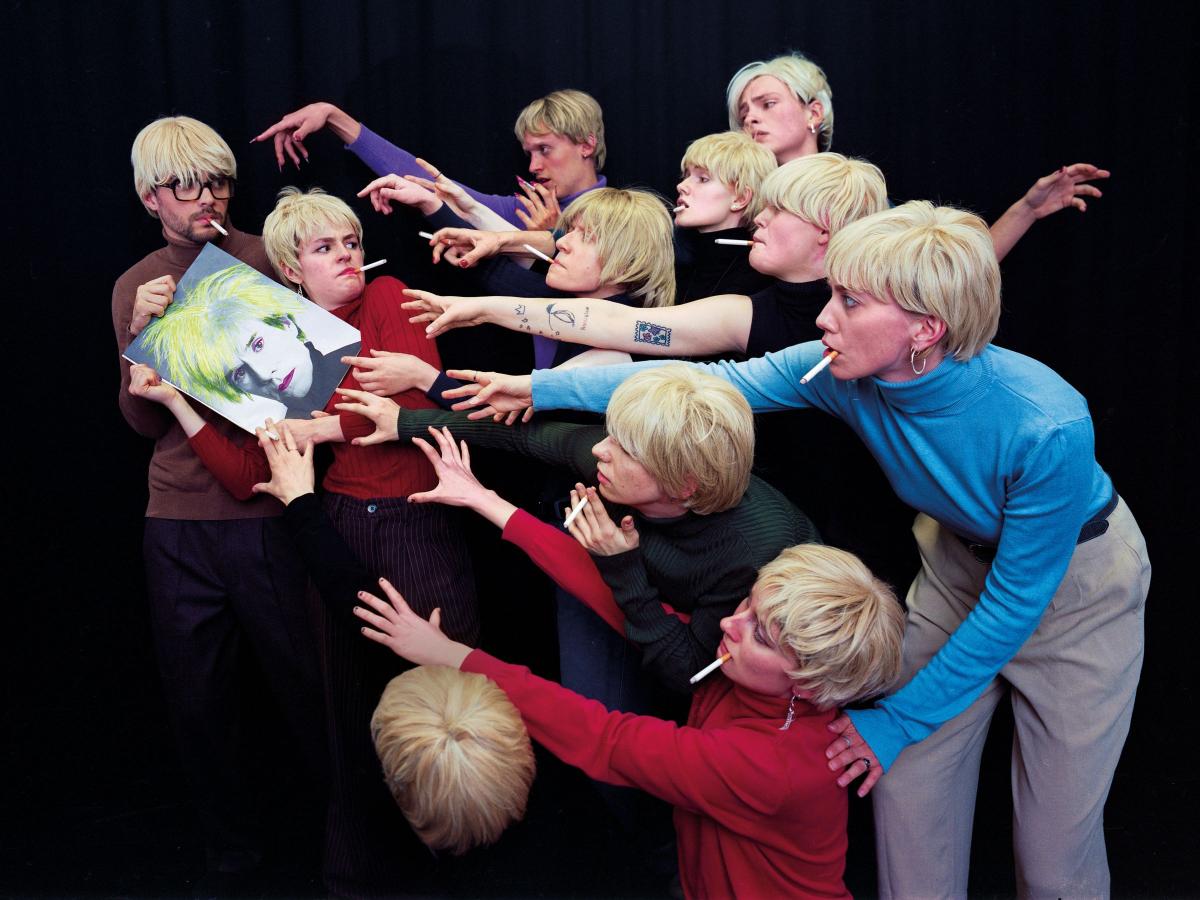 Eine Gruppe junger Menschen mit weißblonden Perücken recken sich nach einem Bild von Andy Warhol
