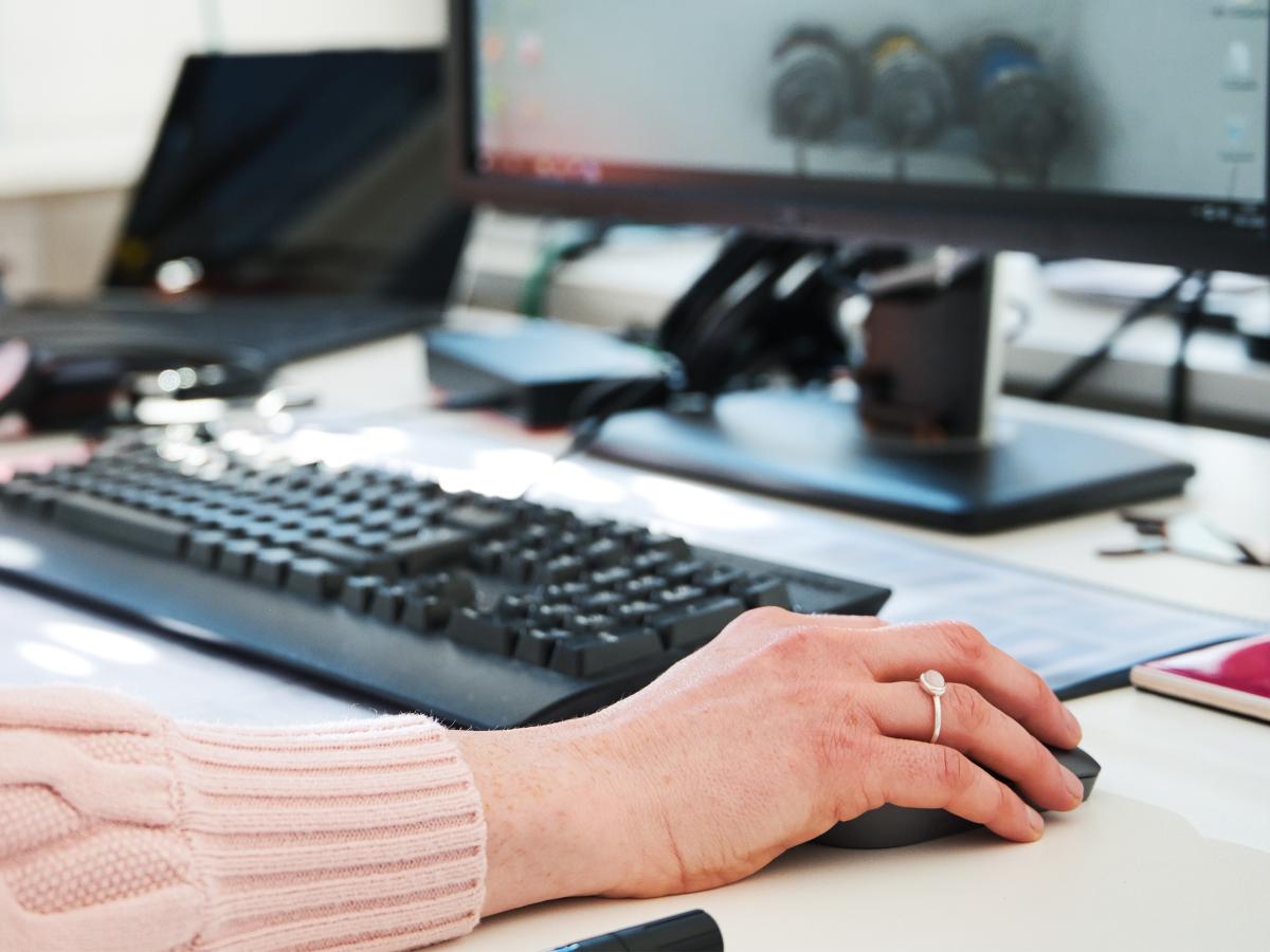 Eine Hand mit bedient die Maus eines Computers. Im Anschnitt ist auch der Bildschirm und die Tastatur zu bedienen. Im Hintergrund steht ein Telefon.