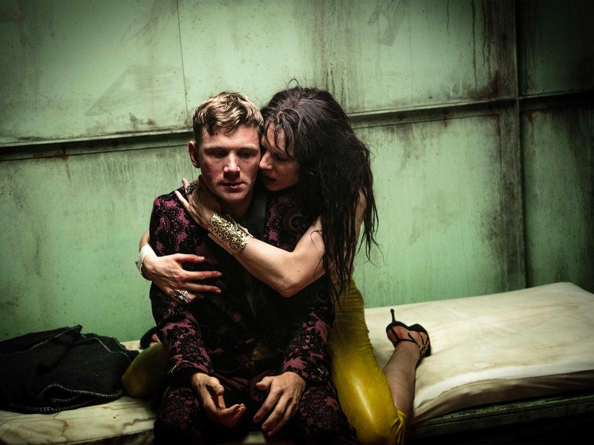 Paul Behren sitzt auf einer schmutzigen gelblichen Matratze vor einer schmutzig-rostigen Wand. Lilith Stangenberg kniet hinter ihm mit gelber Hose, goldenem Armreif und schwarzen Absatz-Schuhen. Sie hat ihre Arme um Paul Behren gelegt und drückt ihr Gesicht an seine linke Wange.