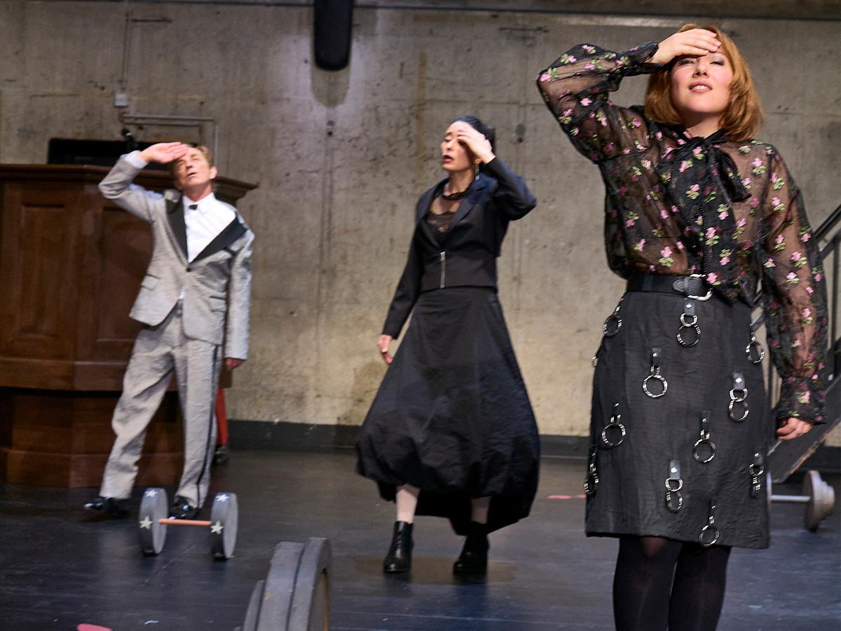 Magne Håvard Brekke, Sasha Rau und Josefine Israel quer im Raum verteilt. Sie haben eine Hand erhoben und fassen sich an den Kopf, die Augen halb geschlossen. Auf den schwarzen Boden sind rote Schuhabdrücke aufgeklebt, eine Kurz- und eine Langhantel liegen herum. Im Hintergrund hängt eine Hantelbank an der Wand in Beton-Optik, die mit einem Spot beleuchtet ist.