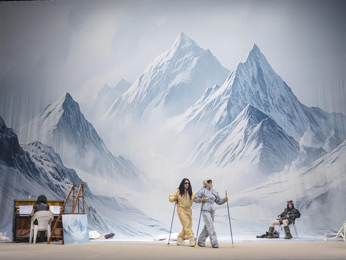 Links sitzt Martin Zamorano am Klavier mit dem Rücken zum Publikum. In der Mitte sind Samuel Weiss und Sachiko Hara, die Skianzüge tragen, Skibrillen auf- und Skistöcke in den Händen haben. Rechts sitzt Sasha Rau auf einem Plastikstuhl, ebenfalls in Skikleidung. Im Hintergrund eine schneebedeckte Berglandschaft. 