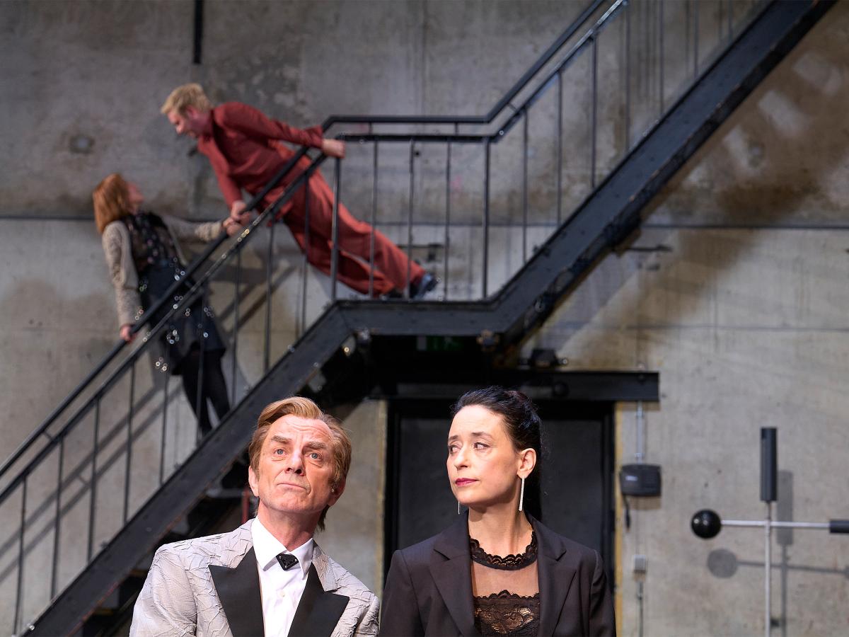 Im Vordergrund Magne Håvard Brekke und Sasha Rau, die ihn anschaut, er schaut geradeaus. Im Hintergrund Josefine Israel und Samuel Weiss auf einer schwarzen Metalltreppe. Josefine Israel hält sich mit beiden Händen am Geländer fest und lehnt sich leicht zurück, während sie nach oben zu Samuel Weiss schaut. Dieser hält sich ebenfalls mit beiden Händen fest und neigt sich mit geradem Körper zu ihr herunter.