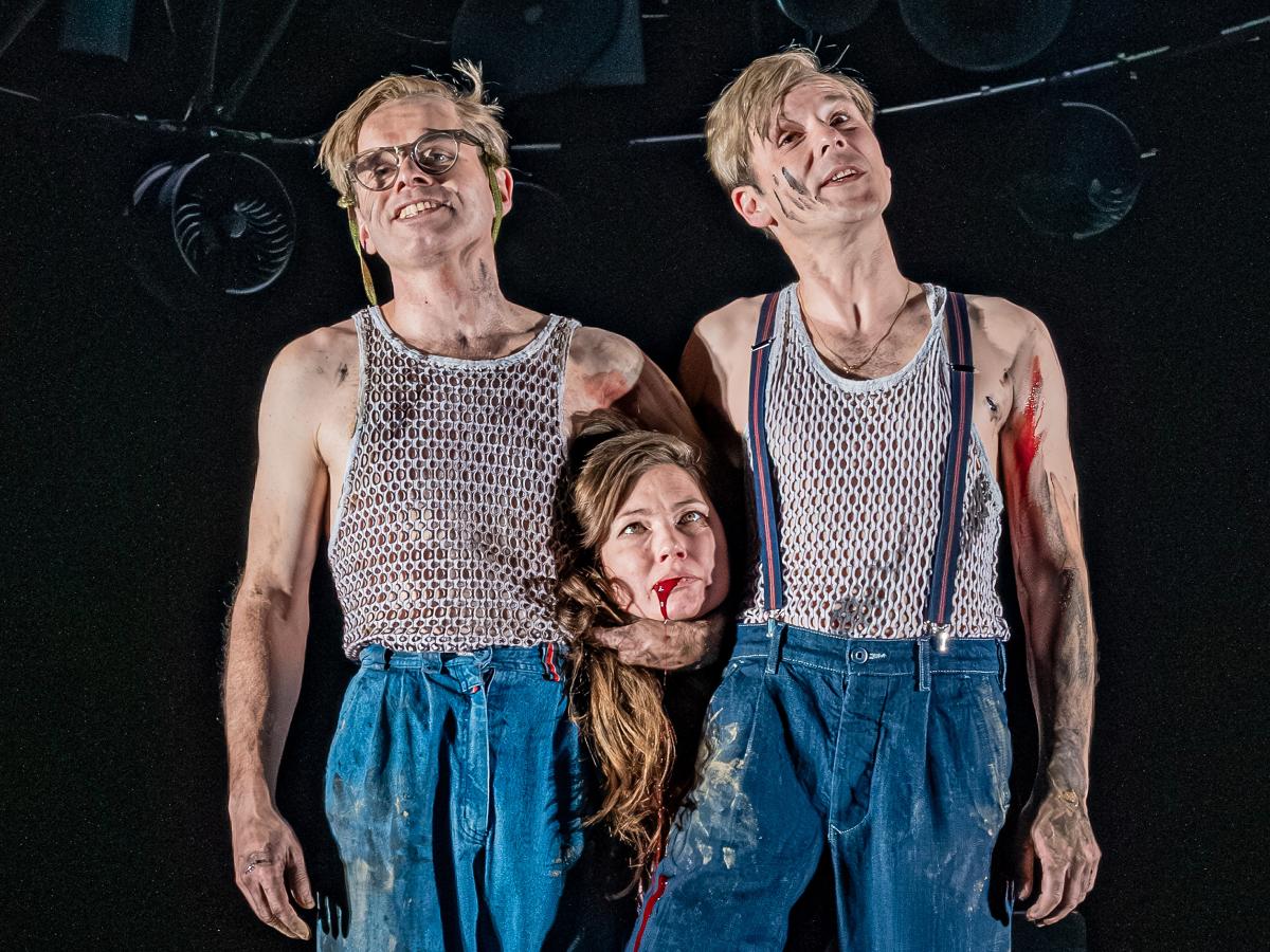 Kristof Van Boven und Nils Kahnwald stehen nebeneinander auf der Bühne. Sie tragen dreck- und blutverschmierte blaue Jeanshosen und graue Unterhemden mit vielen kleinen Löchern. Beide lächeln, unter dem Arm von Kristof Van Boven ist der Kopf von Sabine Molenaar zu sehen, ihr Haar fällt herunter, sie schaut nach oben, aus ihrem Mund läuft Blut. Über ihnen hängt eine große runde Stahlkonstruktion aus vielen unterschiedlich großen alten trichterförmigen Lautsprechern, Scheinwerfern und Ventilatoren. 