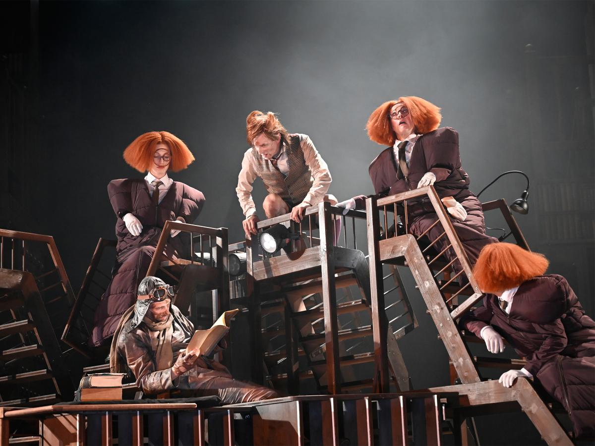 Auf einer Ansammlung von Treppen hockt Jan-Peter Kampwirth und blickt nach unten zu Jan Thümer mit Pilotenmütze, der in einem Manuskript liest. Ebenfalls auf den Treppen sitzen drei Personen mit aufgeplusterten orangenen Perücken. Sie blicken in alle Richtungen.