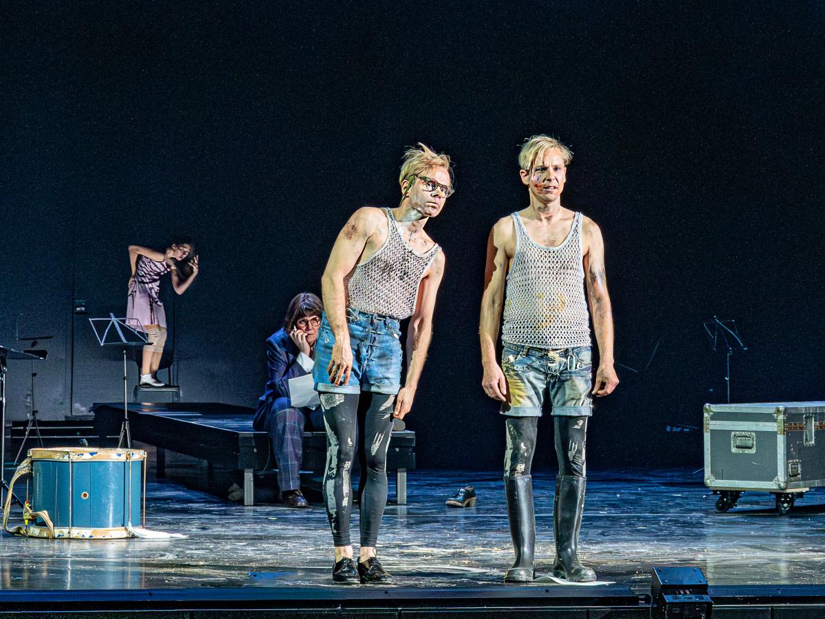 Kristof Van Boven und Nils Kahnwald stehen nebeneinander auf der Bühne. Kristofs Oberkörper neigt sich etwas in Richtung von Nils, die beiden blicken in dieselbe Richtung. Im Hintergrund sitzt Julia Wieninger auf einem Podest und beobachtet die beiden. Auf der Bühne verstreut stehen Notenständer, eine Kiste und eine Trommel. Weiter im Hintergrund tanzt eine Tänzerin.