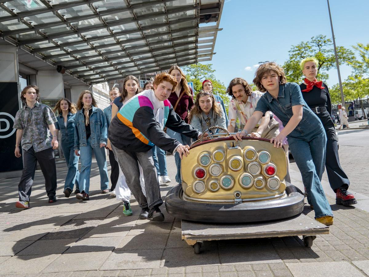 Vier Jugendliche schieben auf einem großen Rollbrett einen bunten Autoscooter vor dem Haupteingang des Schauspielhauses und blicken dabei in die Kamera. Eine Gruppe weiterer Jugendlicher folgt ihnen. Einige lächeln, andere haben einen neutralen Gesichtsausdruck. Sie tragen normale Alltagskleidung.