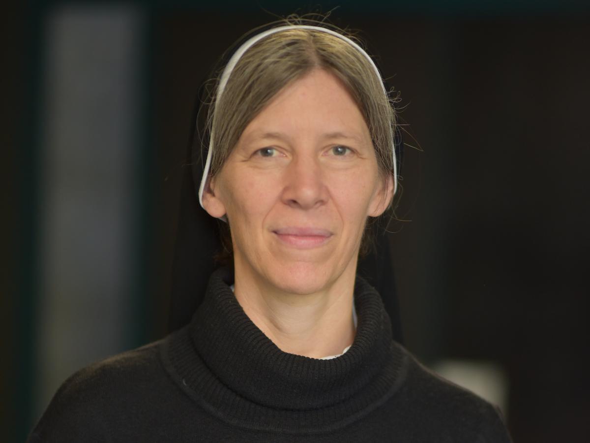 Sr. Britta Müller-Schauenburg CJ steht vor einem dunklen Hintergrund. Sie trägt ein Ordensgewand und schaut in die Kamera.