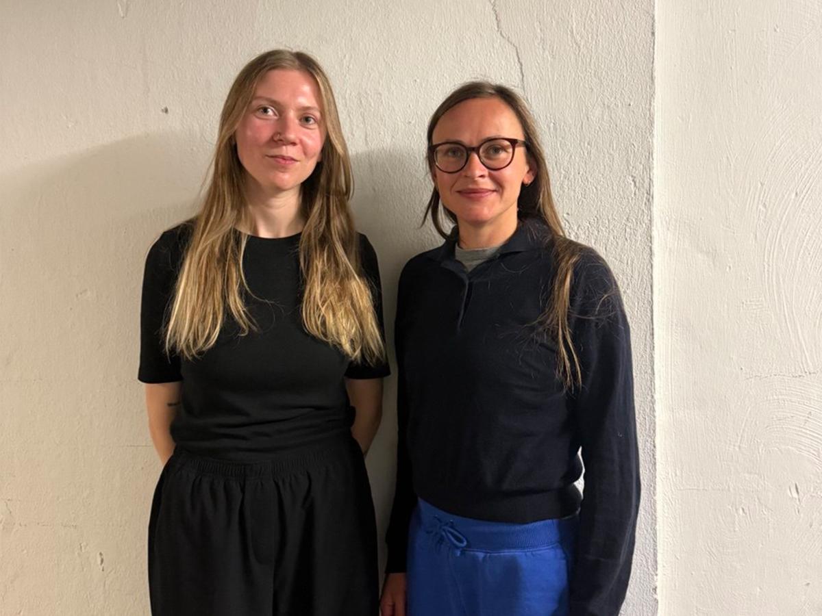 Henni Jörissen und Paula Bühler stehen vor einem neutralen Hintergrund. Henni Jörissen trägt eine dunkle Brille, lange braune Haare, ein schwarzes Oberteil und eine blaue Hose. Paula Bühler trägt lange blonde Haare, ein schwarzes Oberteil und eine schwarze Hose.