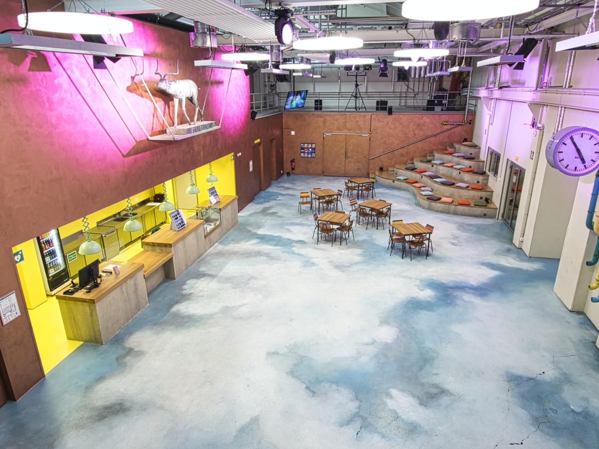 Das Foyer des Junges Schauspielhauses. Buntes Licht, links eine Bar für Getränke, hinten rechts eine Sitzecke. Oben links an der Wand hängt eine hölzerne Kuh.