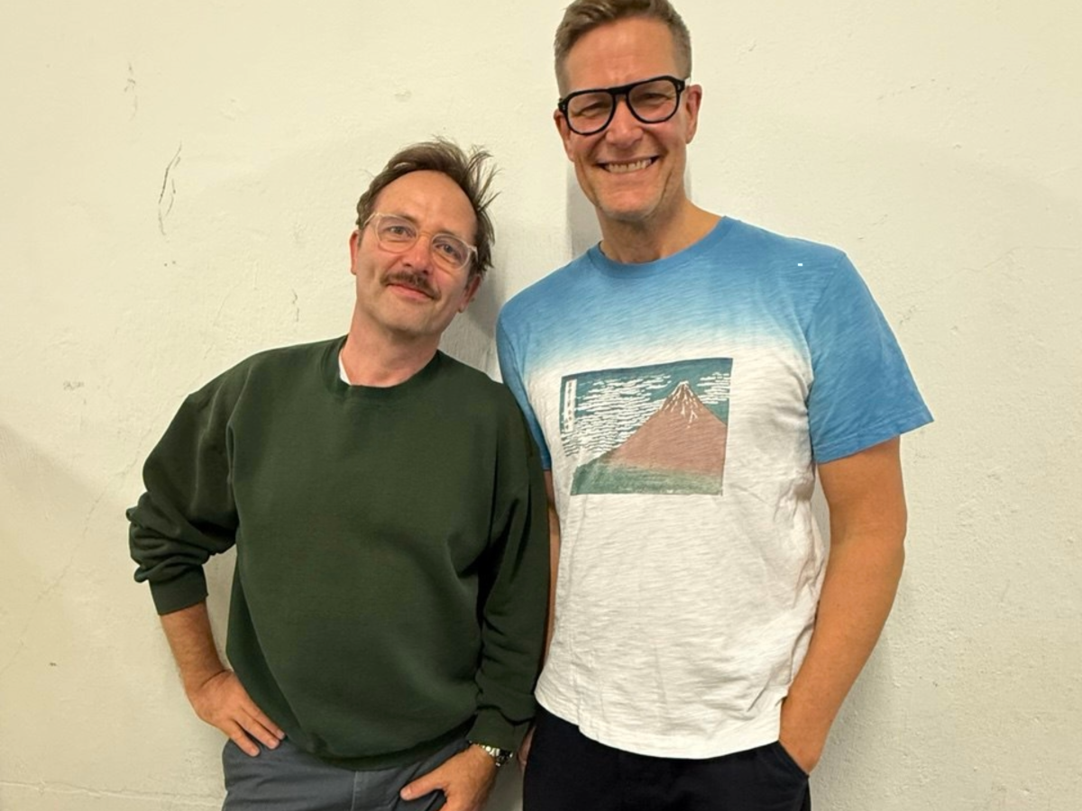 Samuel Weiss und Götz Bühler stehen vor einem neutralen Hintergrund. Samuel Weiss trägt einen grünen Pullover und eine graue Jeans. Götz Bühler trägt eine dunkle Brille und ein blau-weißes Shirt mit Print drauf.