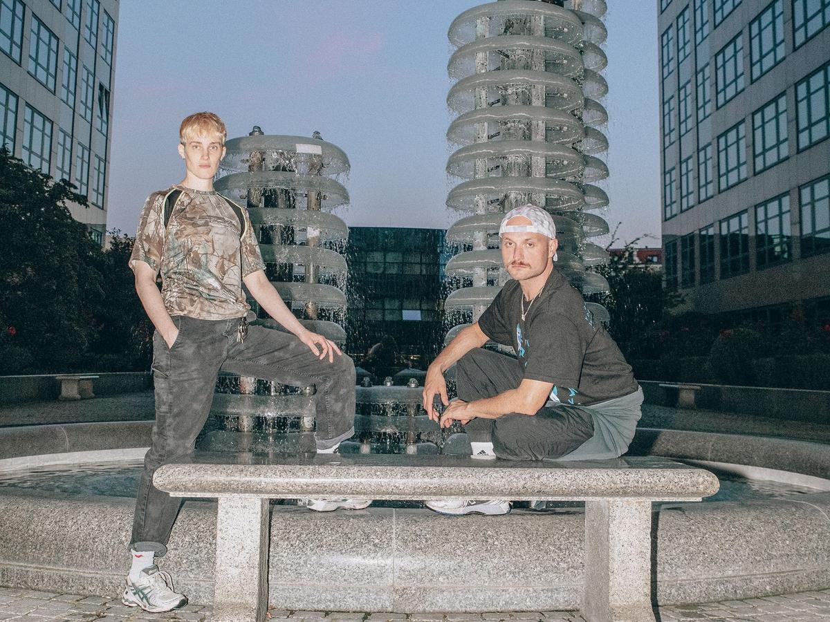 lynn t musiol steht an einem Wasserspiel-Brunnen und hat ein Bein auf dem Rand des Brunnens abgestellt. Neben ihr hockt Marcus Peter Tesch auf dem Rand des Brunnens. Vor den beiden steht eine steinerne Sitzbank.