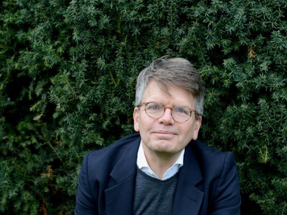 Christoph Möllers sitzt vor einer grünen Nadelhecke. Er trägt eine runde Brille, kurzes, braun-graues Haar, ein weißes Hemd, darüber einen grauen Pullover und ein dunkelblaues Jackett. Er schaut in die Kamera.