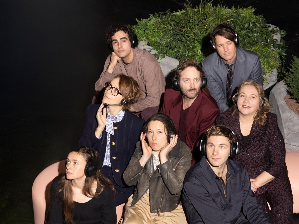 Das Ensemble von „Die Möwe“ sitzt verstreut auf beigen Sitzkissen. Sie tragen alle Overaer-Kopfhörer und blicken in verschiedene Richtungen. Dabei haben sie unterschiedliche Gesichtsausdrücke: Manche lächeln, andere schauen gelangweilt und manche nachdenklich. Im Hintergrund steht ein großer Blumentopf mit gemischten Grünpflanzen.