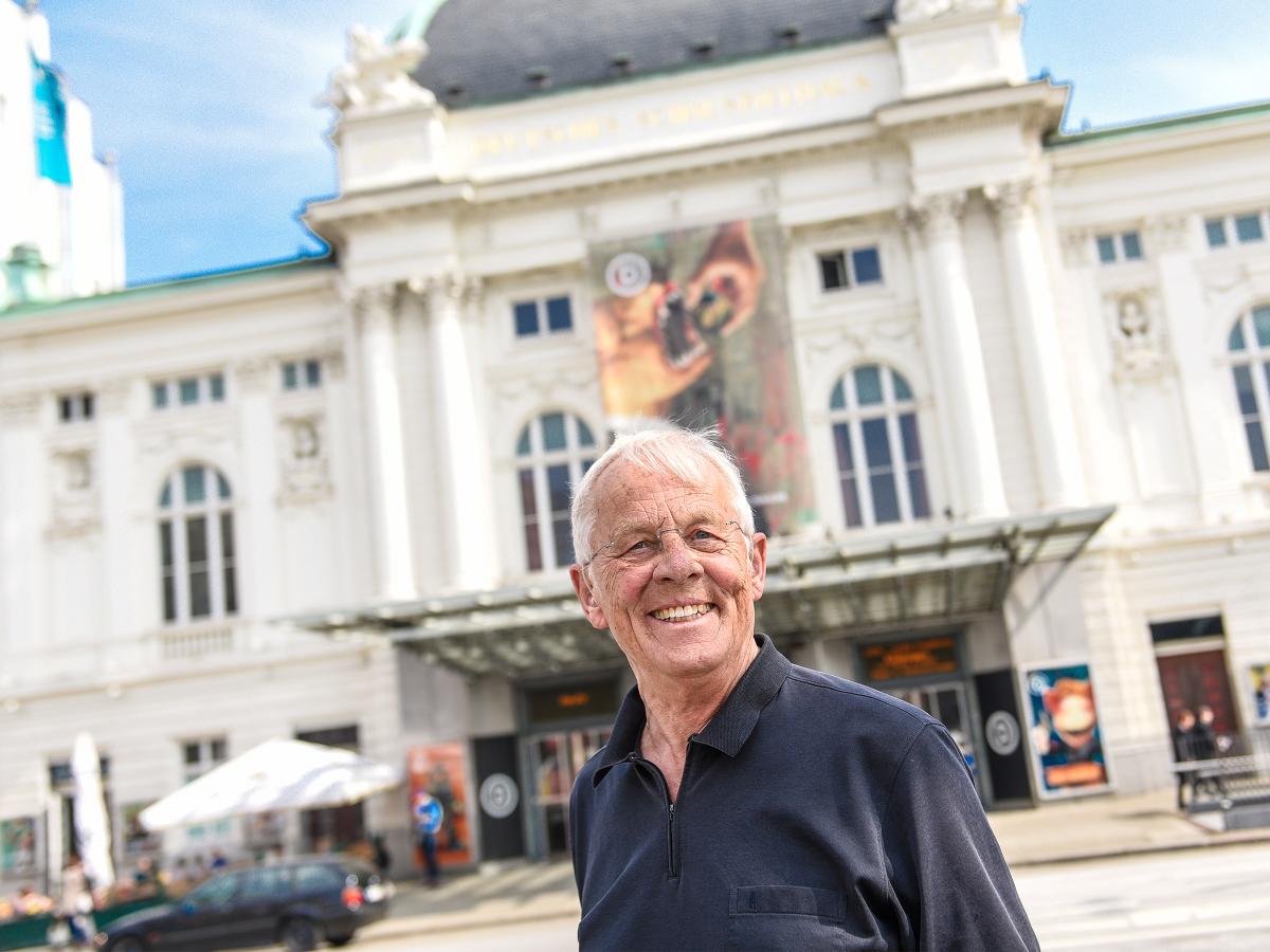 Der Schauspieler Rolf Becker steht draußen vor dem Haupteingang des Deutschen Schauspielhauses. Er trägt ein dunkles Hemd, kurze, weiße Haare und lächelt in die Kamera. Im Hintergrund ist die weiße Fassade des Theaters vor blauem Himmel zu sehen.