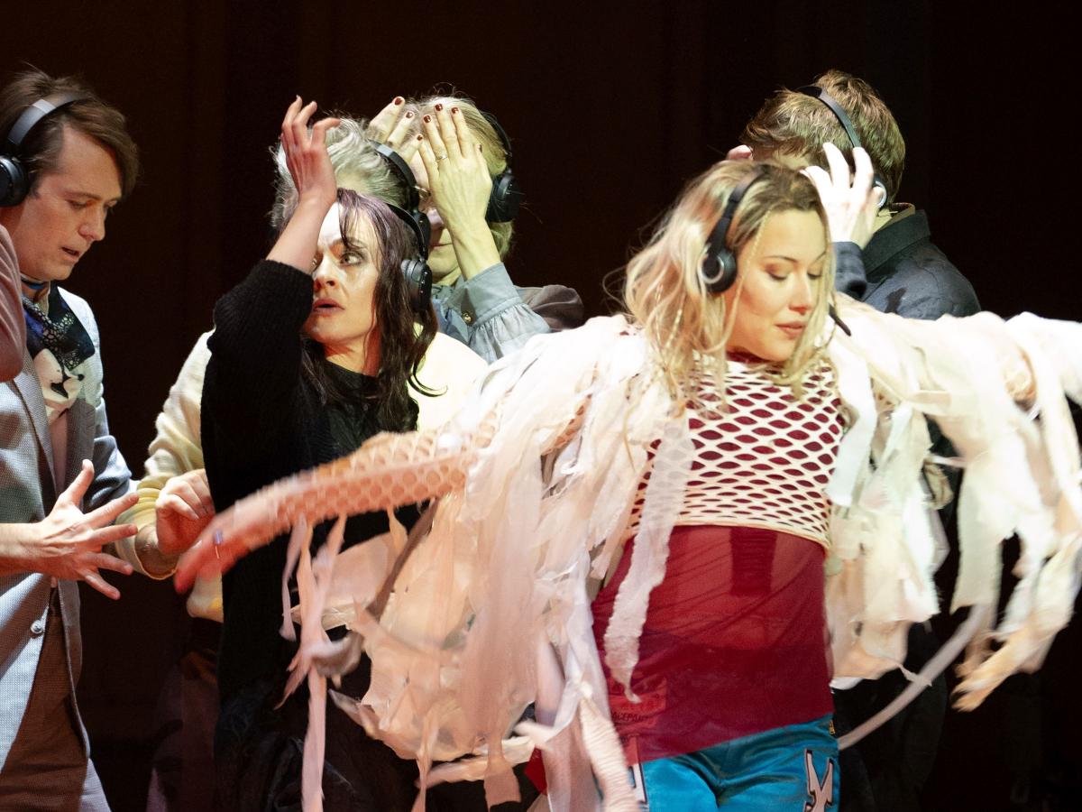 Josefine Israel trägt ein rotes Oberteil und darüber ein weißes Netzoberteil, das an den Armen mit langen und großen Fransen versehen ist. Sie bewegt sich mit ausgestreckten Armen tänzerisch. Im Hintergrund ist das Ensemble zu sehen, sie alle tragen schwarze Kopfhörer auf dem Kopf und bewegen ihre Hände und Arme. 