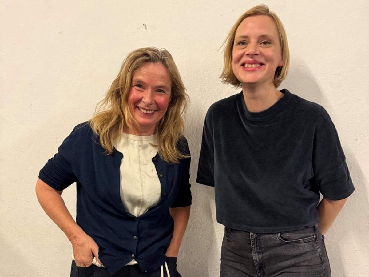 Gewandmeisterin Geseke Brandis und ByteFM-Moderatorin Christa Herdering stehen vor einem neutralen Hintergrund. Geseke Brandis trägt lange blonde Haare, eine weiße Bluse, eine blaue Strickjacke und eine dunkle Hose. Christa Herdering trägt schulterlange hellblonde Haare, ein dunkles Shirt und eine graue Jeans. Sie lächeln in die Kamera. 