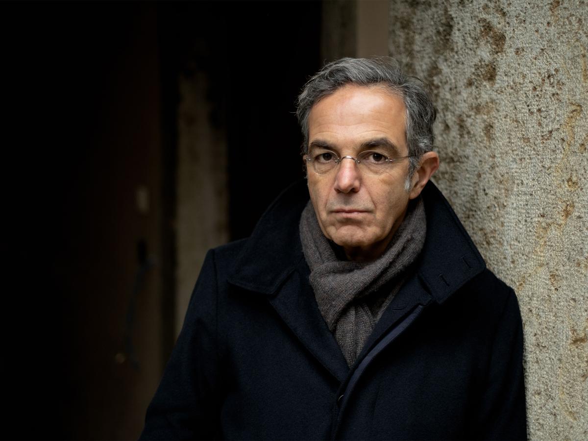 Der Autor Navid Kermani steht vor einer Steinwand. Er trägt einen dunklen Mantel, einen grauen Schal, kurze graue Haare und eine dezente Brille.