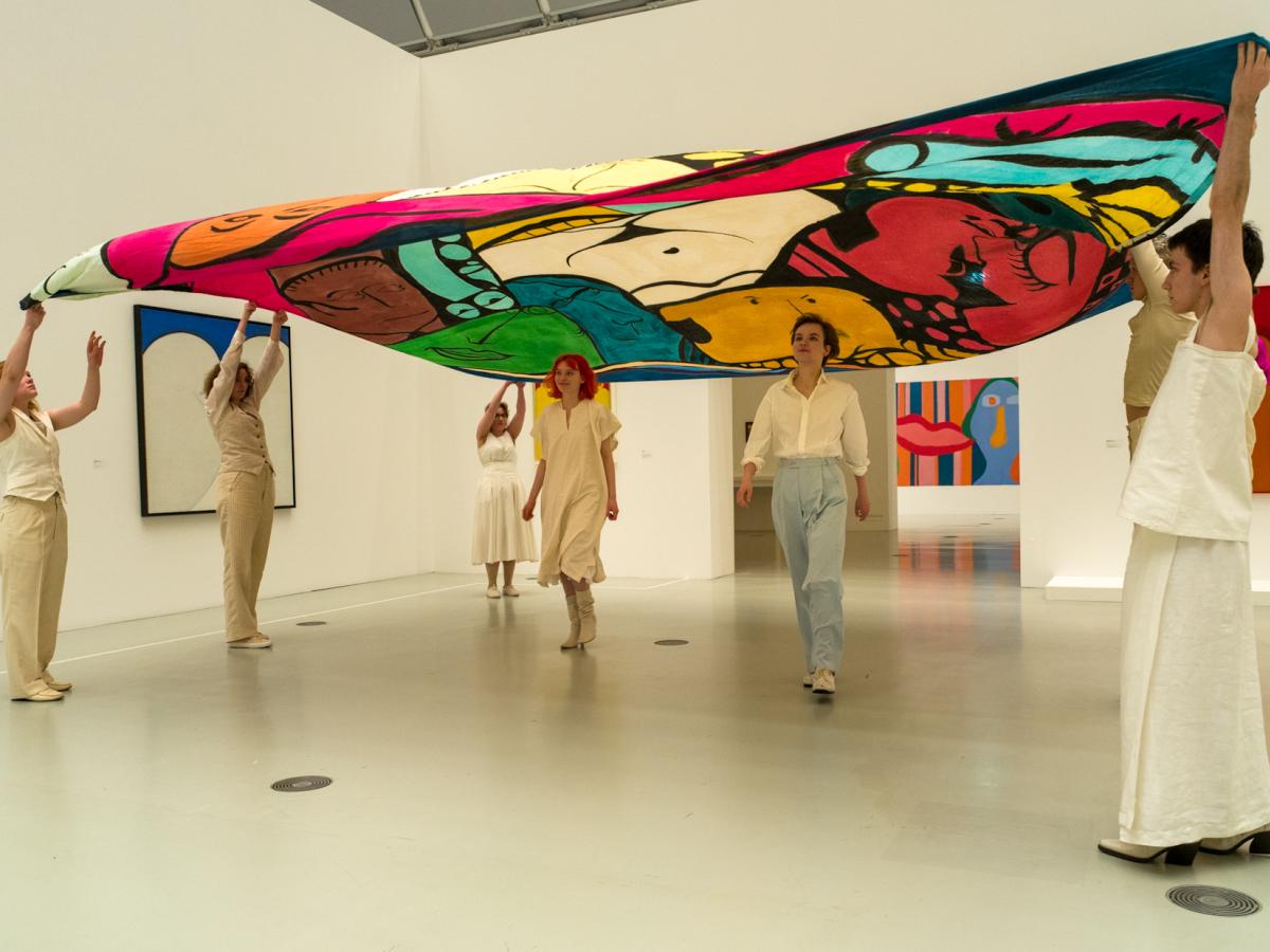 Das Ensemble steht in einem Ram der Deichtorhallen. Sie halten gemeinsam ein buntes Tuch in die Höhe, unter dem zwei Personen entlanglaufen. Alle sind sehr hell gekleidet, in beige und weiß.