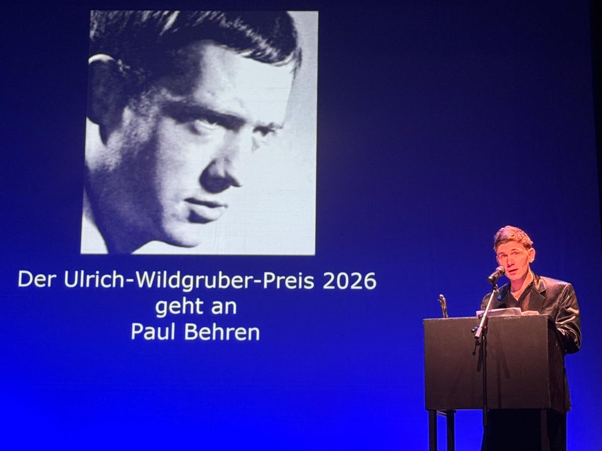 Bühnensituation. Im Hintergrund ist ein blauer Hintergrund zu sehen. Darauf projiziert ist ein Bild des Schauspielers Ulrich Wildgruber. Darunter steht "Der Ulrich-Wildgruber-Preis 2026 geht an Paul Behren". An einem Pult vor dem blauen Hintergrund steht ein schwarzes Pult. Dahinter steht der Schauspieler Paul Behren und spricht in Richtung des Publikums. Er trägt einen schwarzes Satin-Anzug.