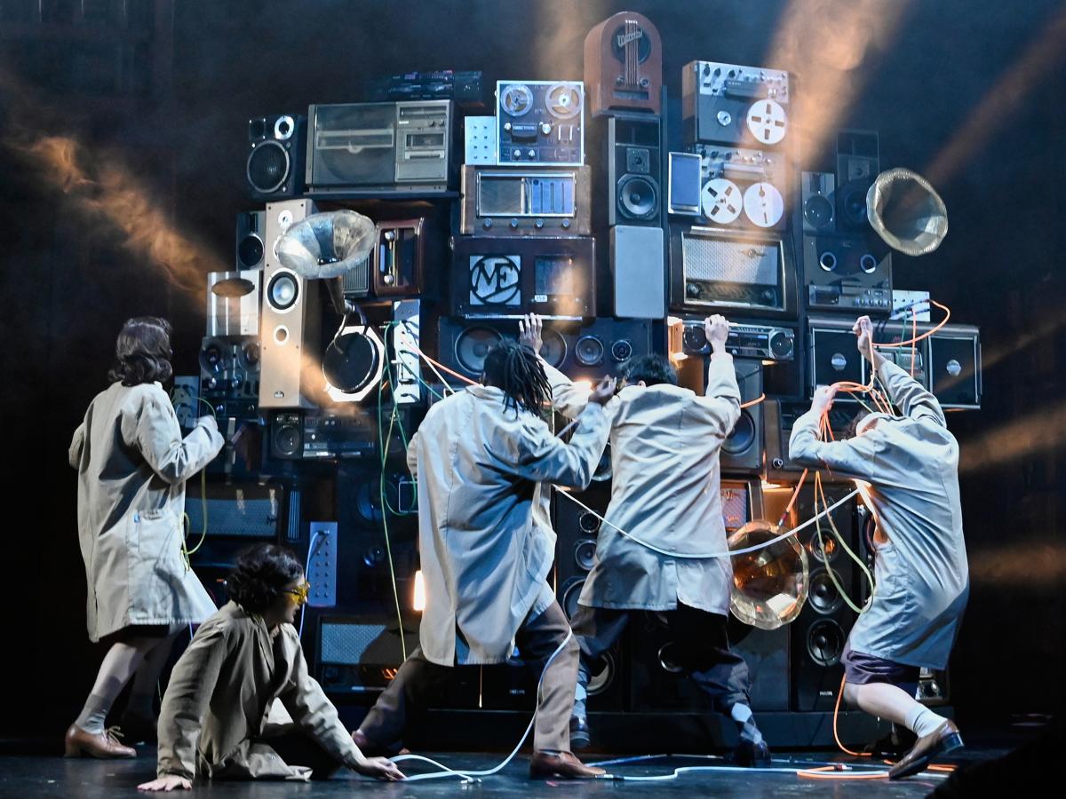 Aus mehreren Stereo- und Musikanlagen, Musikboxen und Grammophonen ist ein großer Gerätehaufen aufgebaut, vor dem fünf Personen stehen, die an den Anlagen rumdrehen.