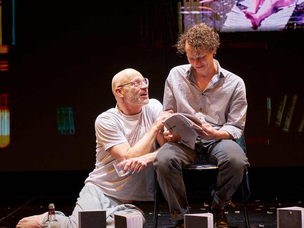 Szenenfoto: Joachim Meyerhoff kniet mit einem Glas in der Hand und lehnt sich an einen Stuhl, auf dem Felix Kammerer sitzt. Kammerer schaut auf ein Heft in seiner Hand und lächelt. Meyerhoff berührt seinen Arm und schaut ihn an, sein Mund ist geöffnet.