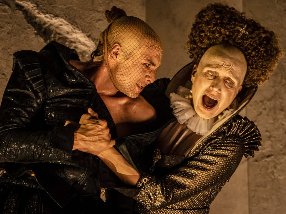 Szenenfoto: Eine Person mit schwarzer Jacke und hautfarbener Netzstrumpfmaske über dem Kopf hält die Hand einer weiteren Person und schaut sie an. Die zweite Person hat ein weiß geschminktes Gesicht und eine hochgesteckte Lockenfrisur mit sehr hoher Stirn. Sie trägt einen Rüschenkragen und eine golden schimmernde Jacke mit Verzierungen. Sie hat den Mund weit geöffnet und schaut zur Seite.