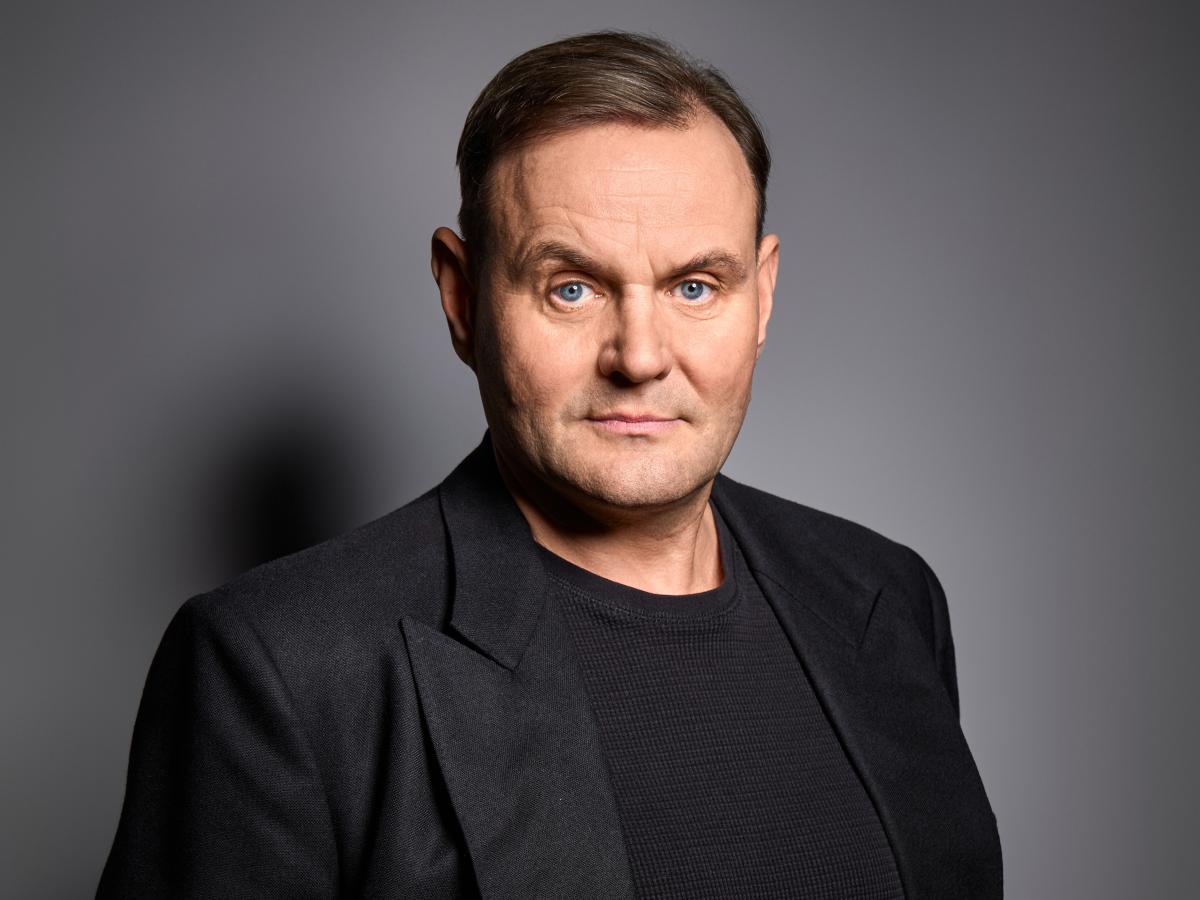 Devid Striesow steht vor einem grauen Hintergrund. Er trägt ein schwarzes T-Shirt, ein schwarzes Jackett und kurze blonde Haare.