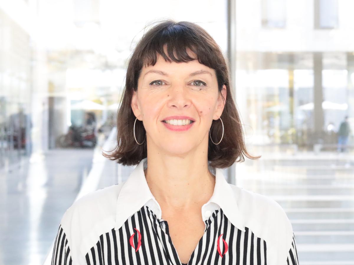 Prof. Dr. Elke Schwarz steht in einem Innenraum vor einer großen Fensterfront. Sie trägt schulterlange dunkle Haare, silberne Ohrringe und eine schwarz-weiß gestreifte Bluse mit roten Details.