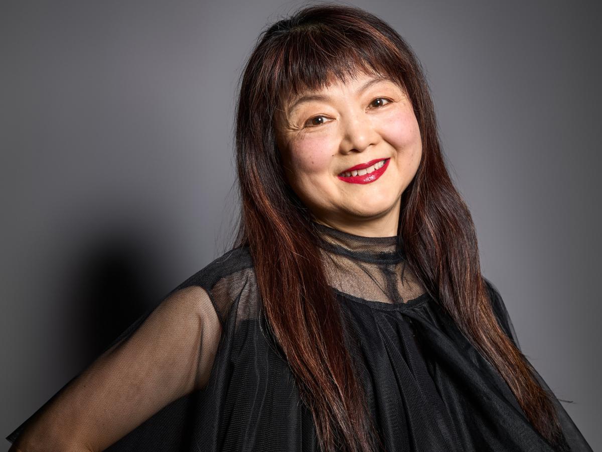 Sachiko Hara steht vor einem neutralen Hintergrund. Sie schaut in die Kamera und lächelt. Sie trägt ein schwarzes, an den Ärmeln leicht transparentes Oberteil, lange dunkelbraune Haare und roten Lippenstift.