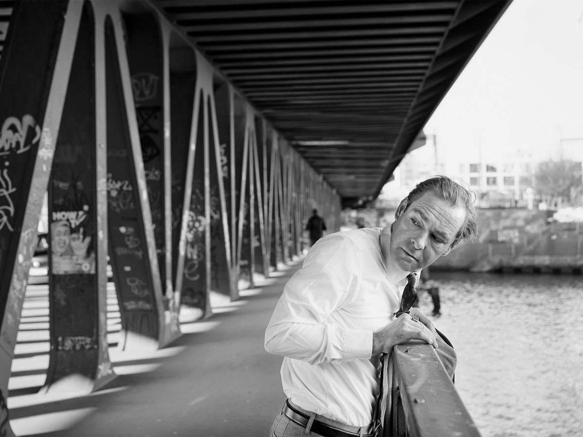 Außenansicht. Schwarz-Weiß-Bild. Mirco Kreibich steht leicht über ein Geländer gebeugt auf einer Brücke über einem Fluss. Links von ihm sind die Brückenaufbauten im Bild. Mirco dreht sich leicht über seine rechte Schulter und schaut hinter sich. Er trägt ein weißes Hemd, eine Krawatte, eine helle Hose mit Gürtel und einen großen Ring am rechten Zeigefinger. Seine Jacke hat er über das Geländer gelegt. Rechts im Bild ist das Wasser des Flusses im Bild.