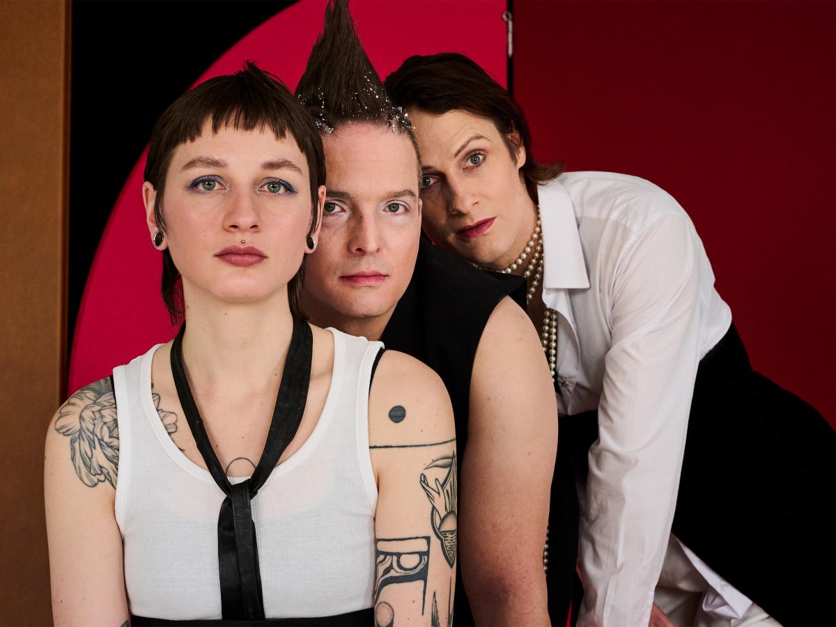 Drei Personen stehen leicht versetzt hintereinander. Der Hintergrund ist farbig, rot und schwarz. Alle drei gucken in die Kamera. Die Person ganz vorne trägt ein weißes Tanktop, eine schwarze Krawatte und kurze dunkle Haare. Die Person in der Mitte trägt ein schwarzes ärmelloses Oberteil und hochgegelte Haare, die glitzern. Die Person ganz hinten trägt eine weiße Bluse, einen schwarzen Rock und viele Perlenketten. Sie hat schulterlange dunkelbraune Haare.