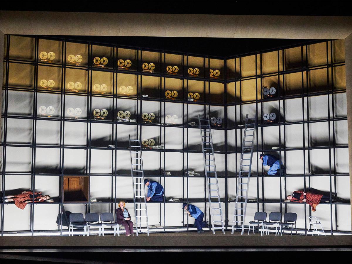 Bühnensituation: Über das Bühnenbild erstreckt sich ein Metallgestell, welches so angeordnet ist, dass sich regalartige Fächer bilden. In den oberen Fächern sind mehrere Tonbandgeräte zu sehen. Etwas weiter unten knien verteilt drei Männer in einzelnen Regalfächern.  Alberta von Polnitz und Julia Wieninger liegen jeweils links und rechts in Fächern, Ilse Ritter sitzt auf einem Stuhl in einer Stuhlreihe.