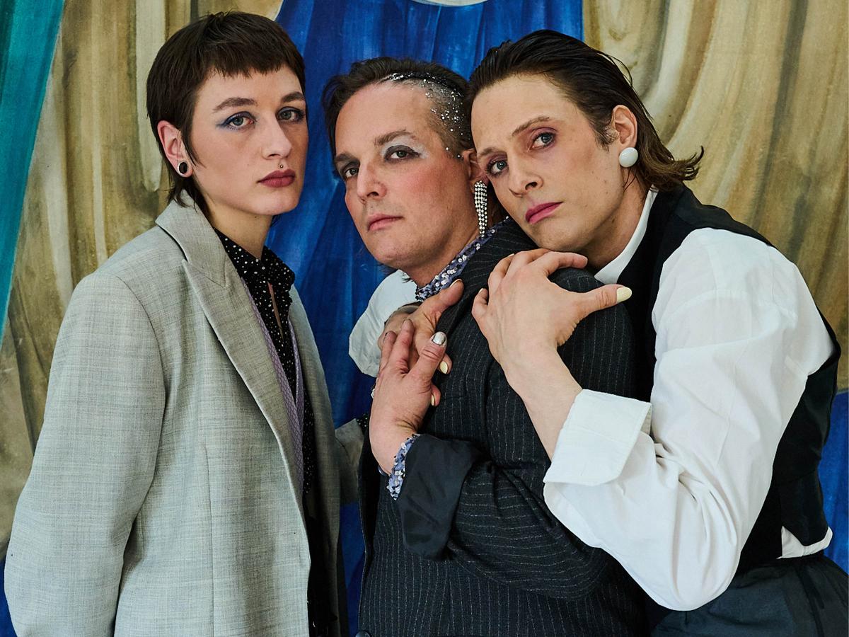 Juno Peter, Micha Krause und Less Brandes-Hell stehen eng nebeneinander und schauen in die Kamera. Less hat ihren Kopf und die Arme um Michas Schultern gelegt, Micha berührt ihre Hand. Sie stehen vor einem gemalten gelben Vorhang.