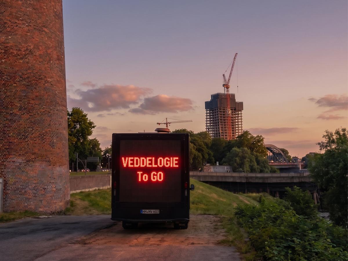 Außenansicht. Links im Bild ist ein großes rotes Backsteingebäude zu erkennen. Davor, auf einem rauen Betonuntergrund, parkt ein schwarzer LKW mit einer großen Leuchtschrift auf der Rückseite. Dort steht: VDDLTOPOLY to GO. Im Hintergrund ist eine Flussumgebung des Hamburger Hafens mit einer Brücke und Autos zu sehen. Ganz im Hintergrund ragt der unfertige bau des Elbtowers mit zwei großen Kränen in den Himmel.