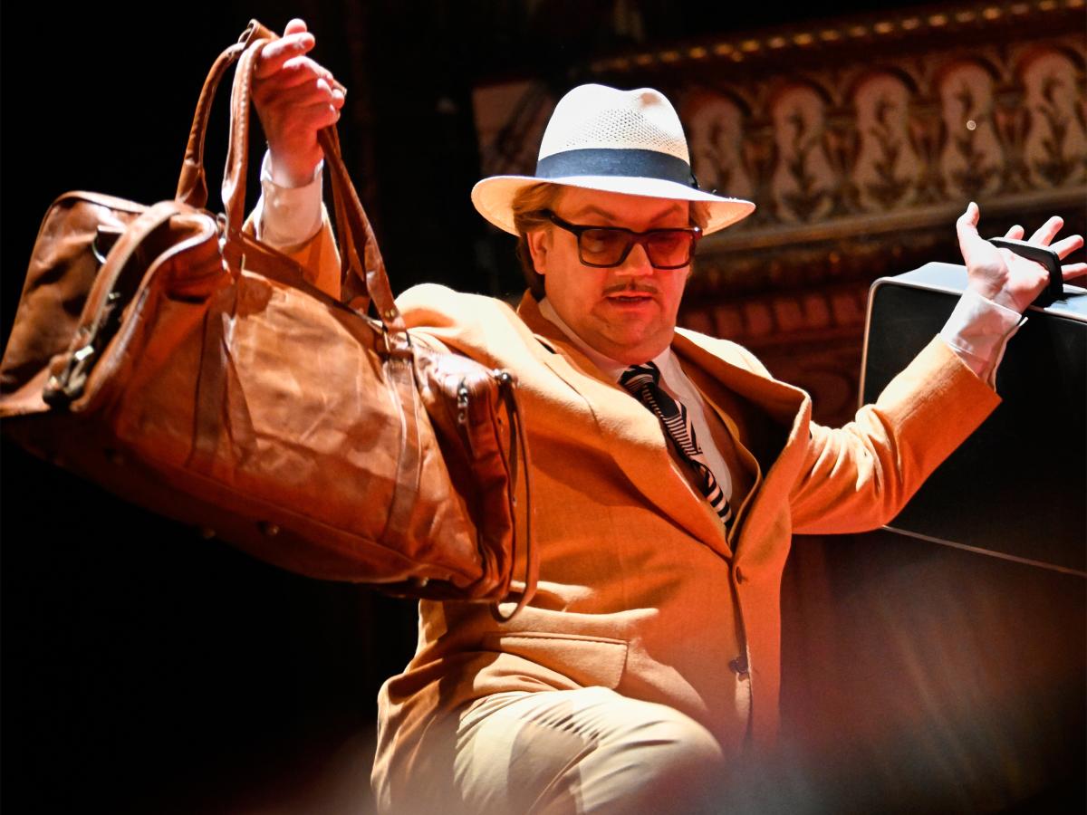 Schauspieler Charly Hübner auf einer Theaterbühne in einem beigen Jackett, gestreifter Krawatte, weißem Fedora-Hut und auffälliger Sonnenbrille mit Schnurrbart. Er hält mit ausgestrecktem Arm eine große braune Ledertasche in die Höhe, während er mit der anderen Hand einen schwarzen Koffer balanciert. Die warme Bühnenbeleuchtung taucht die Szene in ein goldenes Licht.