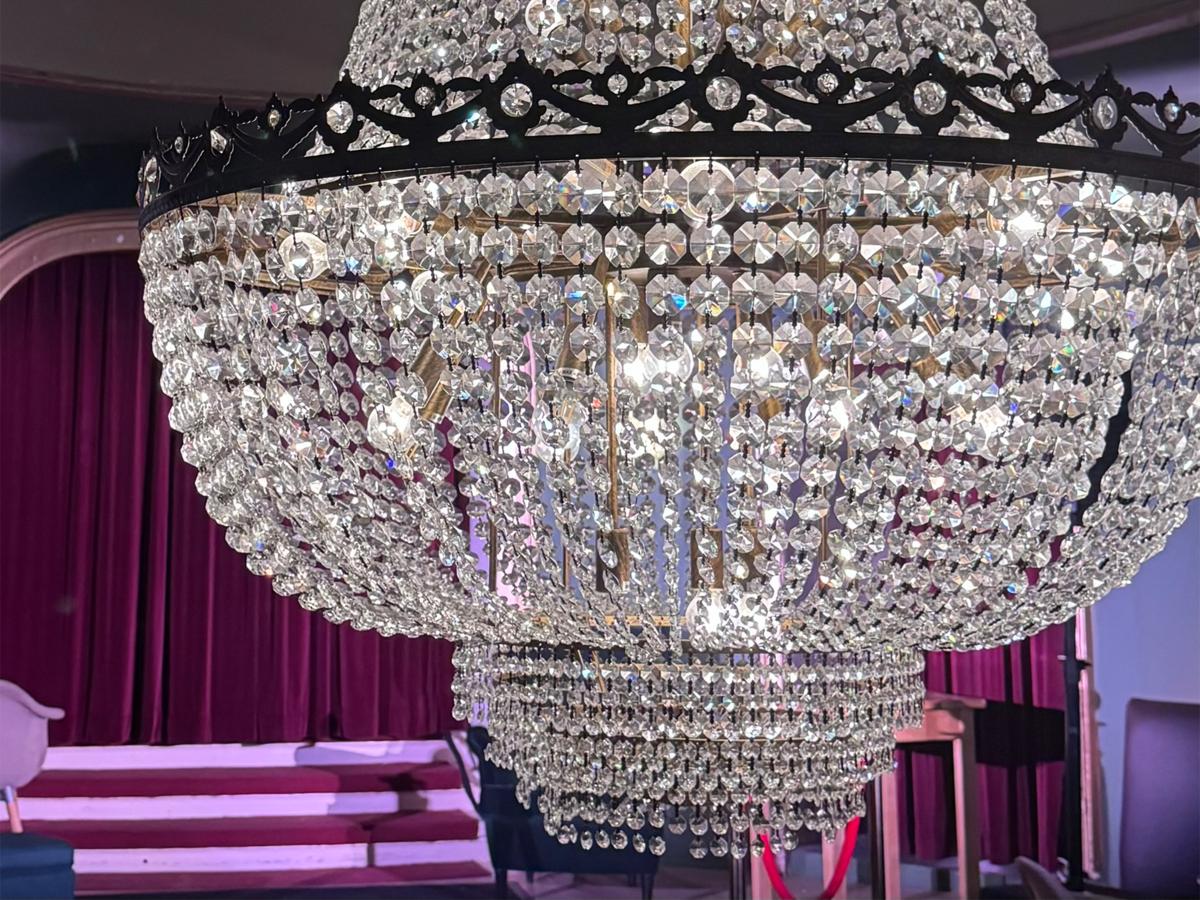 Zu sehen ist eine Großaufnahme des Kronleuchters der Kantine im Schauspielhaus in Hamburg. Hinter dem Leuchter sieht man ein paar Stufen, eine kleine Bühne und einen roten Samtvorhang.