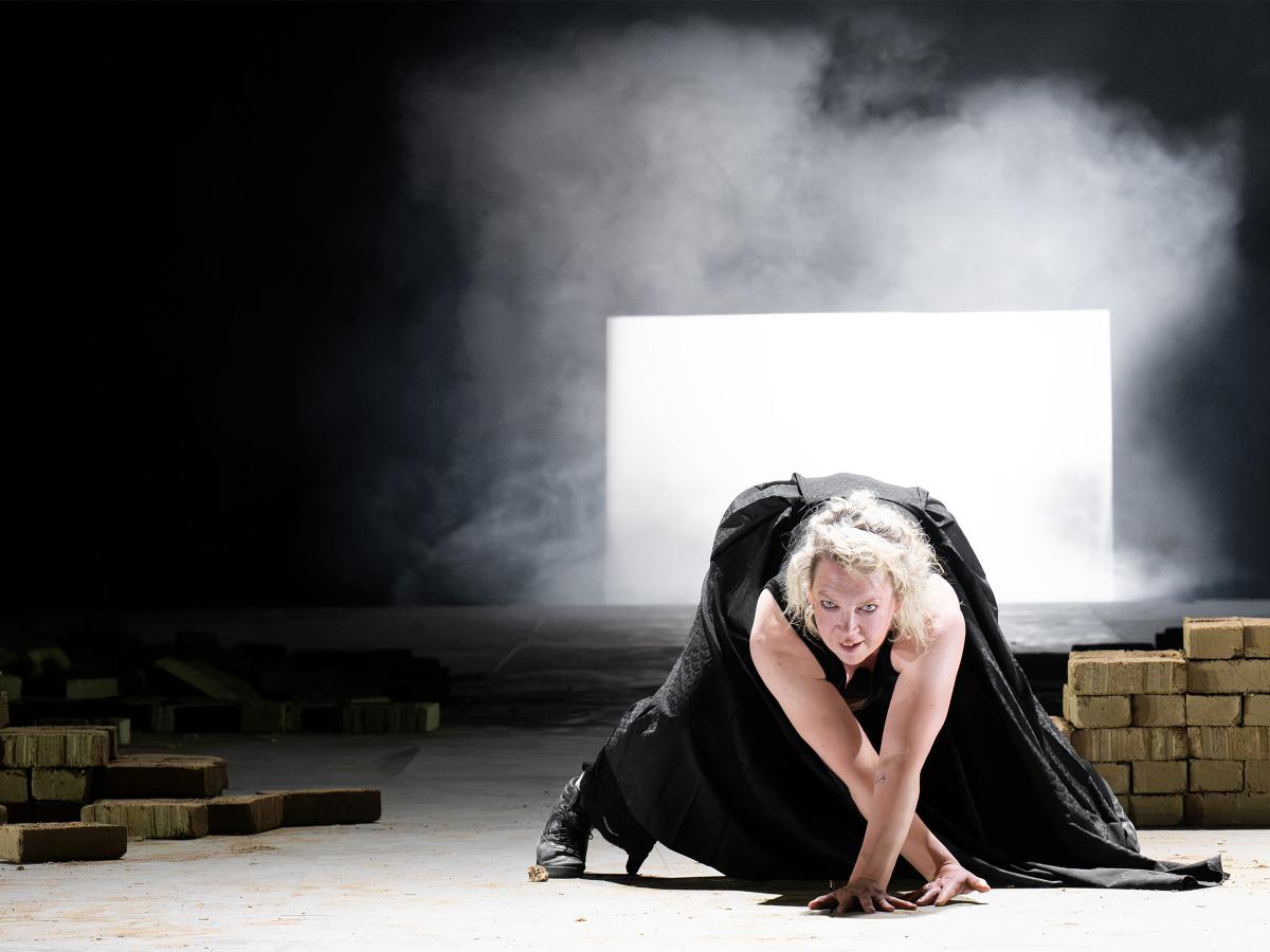 Ankündigungsfoto »Fremde Sonne II: Medea in Korinth«; Lina Beckmann in schwarzem Gewand kauert auf dem Boden der Bühne und stützt sich mit einer Hand ab, den Blick intensiv nach vorne gerichtet. Im Hintergrund ein gleißend helles Rechteck, umgeben von Nebel und Dunkel. Rechts und links verteilt gestapelte Ziegelsteine. Die Szene strahlt Bedrohung und äußerste Anspannung aus.