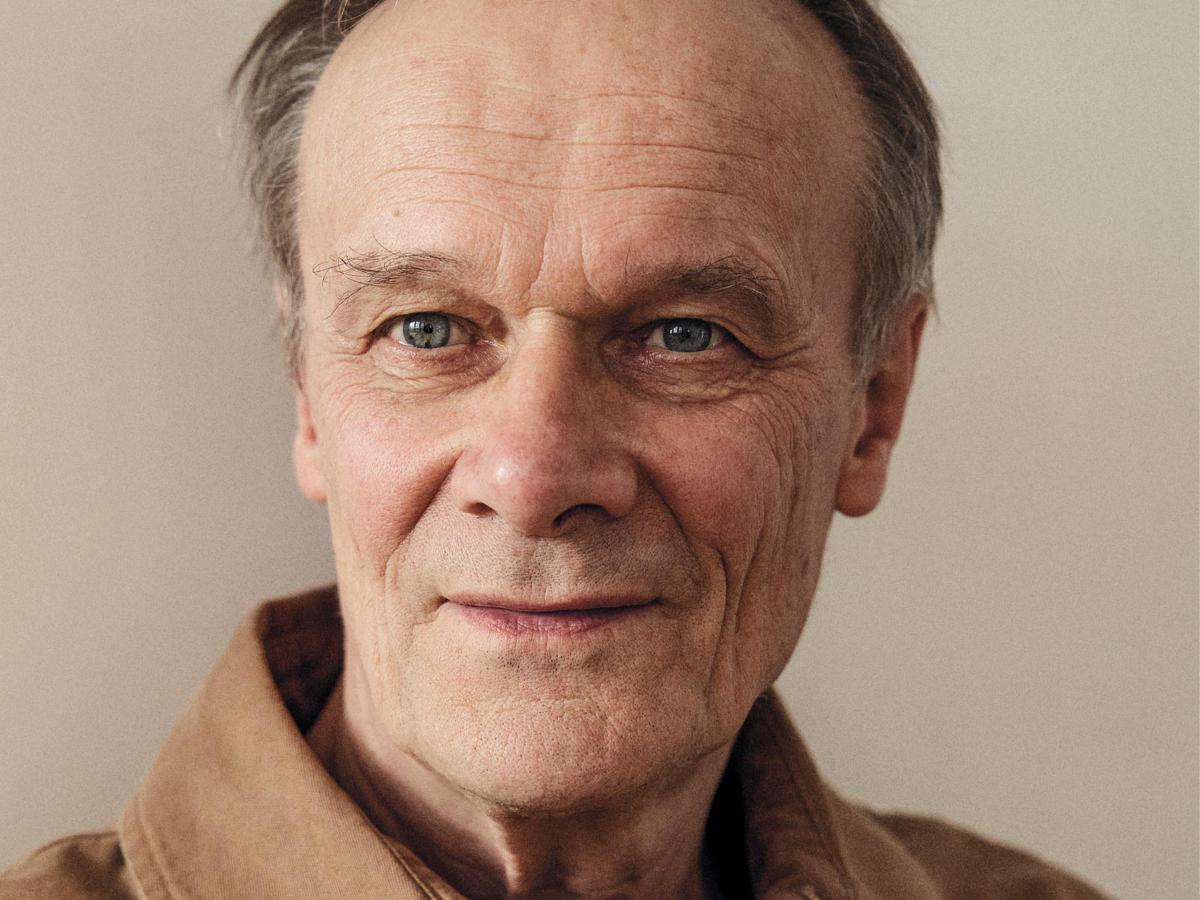 Nahaufnahme von Schauspieler und Autor Edgar Selge. Er trägt kurze graue Haare und ein helles Hemd. Er blickt leicht an der Kamera vorbei und lächelt leicht vor einem neutralen, hellen Hintergrund.