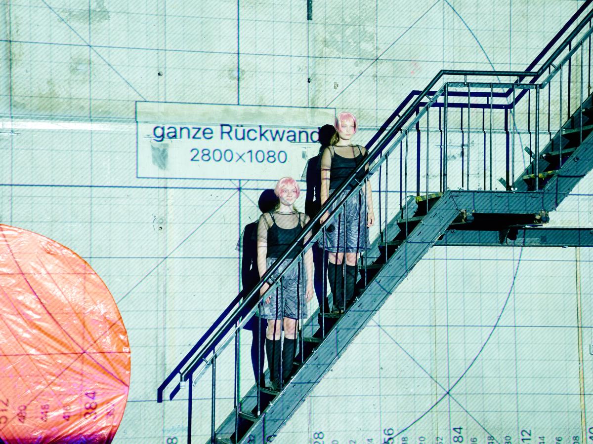 Zwei Personen mit kurzen rosa Haaren stehen auf einer Metalltreppe in einer hell beleuchteten, technisch wirkenden Kulisse. Beide tragen schwarze Oberteile, graue Shorts und schwarze kniehohe Stiefel. Im Hintergrund sind Rasterlinien und eine Beschriftung mit „ganze Rückwand 2800×1080“ zu sehen. Links ragt ein großes orangefarbenes, rundes Objekt ins Bild.