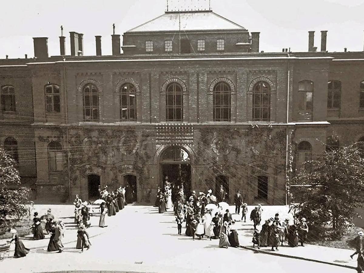 Historische Schwarzweißaufnahme des Universitätsklinikums Hamburg-Eppendorf (UKE). Vor einem repräsentativen Klinikgebäude mit großen Rundbogenfenstern stehen und gehen zahlreiche Menschen, darunter viele Frauen und Kinder. Die Aufnahme zeigt das UKE in historischer Zeit.